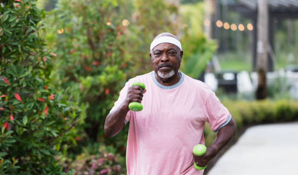 Best Exercises For The Brain A senior African-American man in his 70s exercising outdoors. He is power walking or jogging, with dumbbells in his hands. He is looking at the camera with a serious expression.